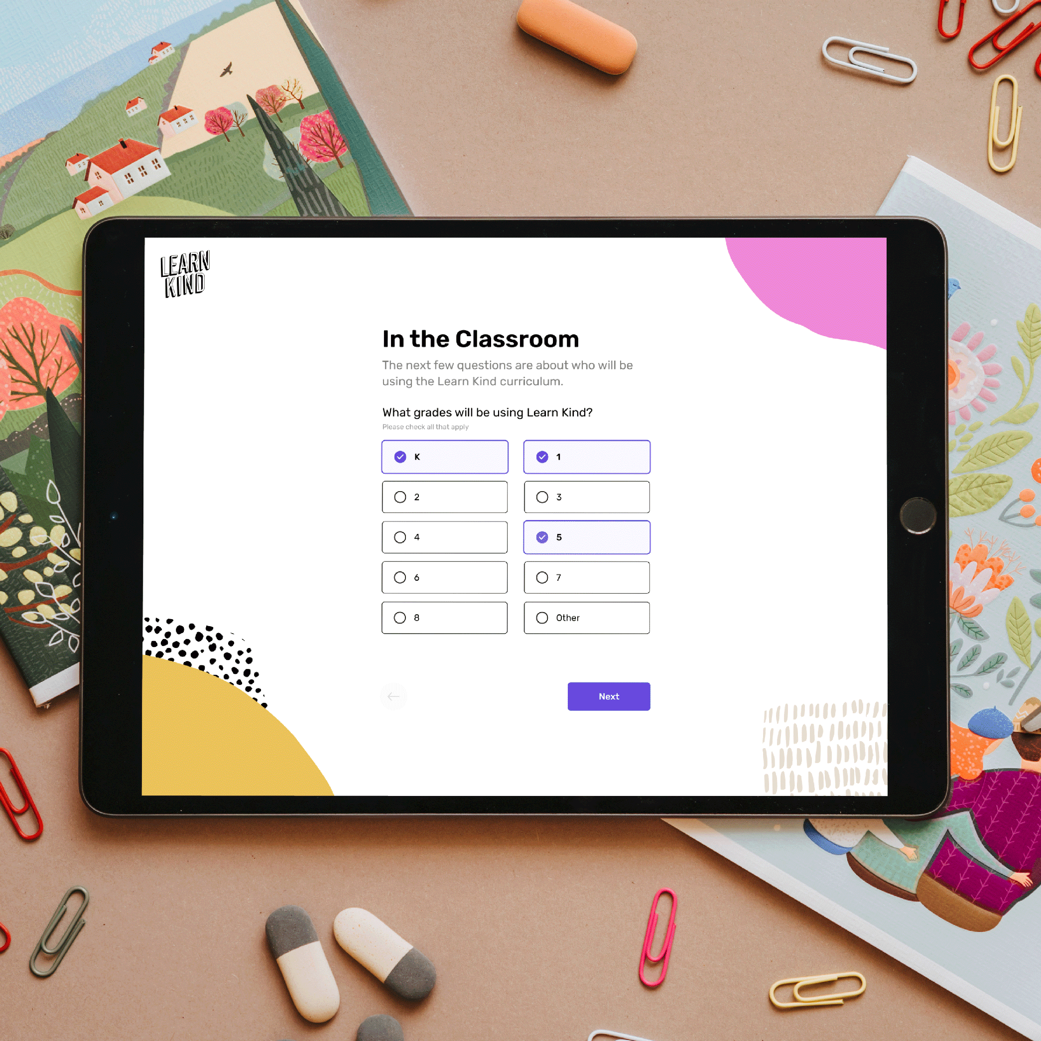 Tablet displaying Learn Kind curriculum selection screen on a desk with colorful paperclips and erasers.